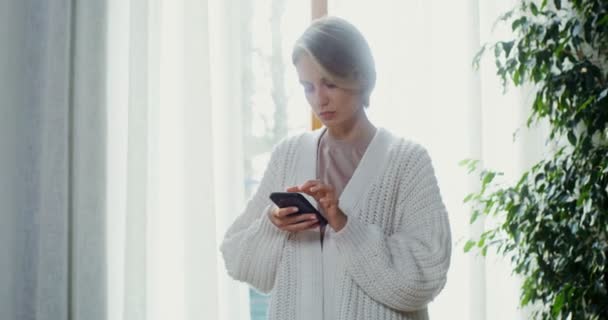 Mujer joven utiliza el teléfono, caminando por la habitación en el interior del hogar moderno — Vídeos de Stock