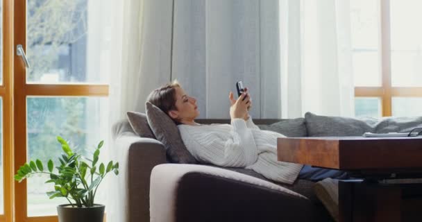Νεαρή γυναίκα χρησιμοποιεί το τηλέφωνο, ενώ βρίσκεται στον καναπέ στο σύγχρονο εσωτερικό του σπιτιού — Αρχείο Βίντεο