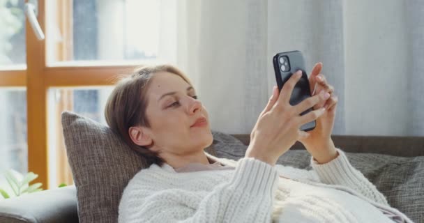 Jovem mulher usa o telefone enquanto deitado no sofá no interior da casa moderna — Vídeo de Stock