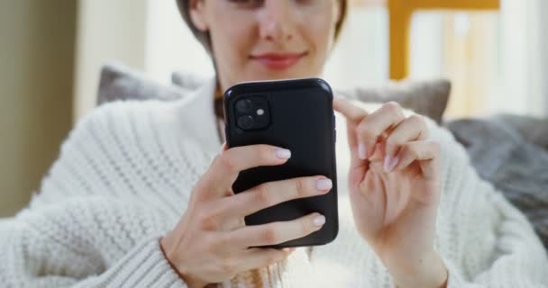 Mujer joven utiliza el teléfono mientras está acostado en el sofá en el interior del hogar moderno — Vídeo de stock