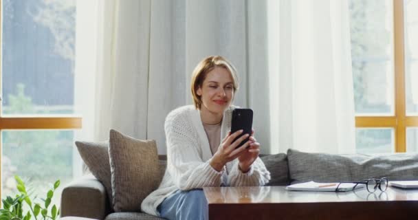Jovem conversando por vídeo chat em um telefone celular enquanto se senta em um sofá — Vídeo de Stock