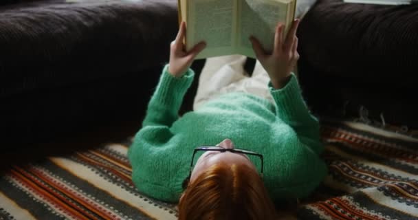 Ένα κορίτσι διαβάζει ένα βιβλίο με ξαπλωμένα στο πάτωμα και αναπαυτικά πόδια στον καναπέ. — Αρχείο Βίντεο