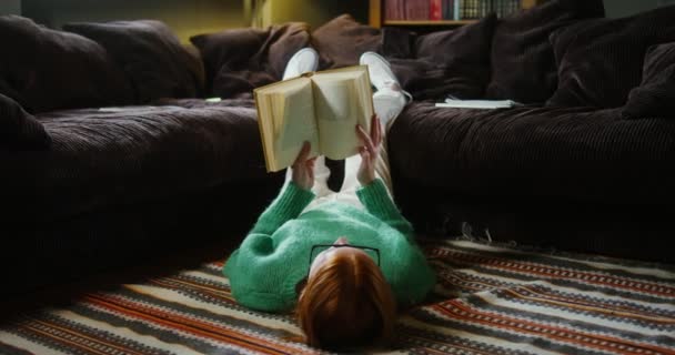 Uma menina lendo um livro com deitado no chão e descansando pernas no sofá — Vídeo de Stock