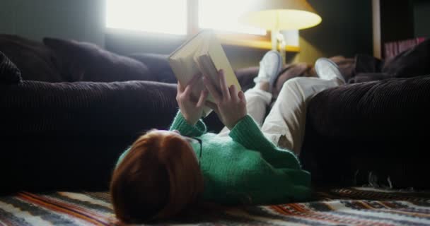 Una chica leyendo un libro tumbada en el suelo y descansando piernas en el sofá — Vídeo de stock