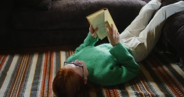A girl tired of reading lays down a book while lying on floor in home interior — Stock Video