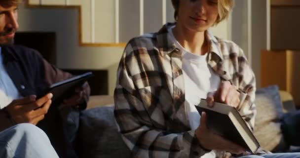 A woman is taking apart a box of books while sitting on the couch next to man — Stock Video