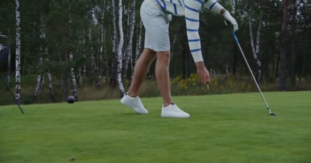 Un hombre joven clava el titular de la pelota de golf en el suelo y coloca una pelota de golf en él — Vídeo de stock