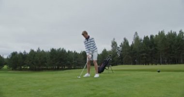 Genç bir adam golf sopasıyla topa vuruyor ve uzağa bakıyor.