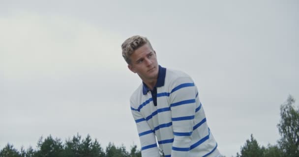 Un joven se prepara para golpear la pelota con un palo de golf, mirando a la distancia — Vídeo de stock