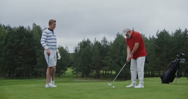 Młody mężczyzna wygląda jak starszy mężczyzna uderza piłkę kijem golfowym. — Wideo stockowe