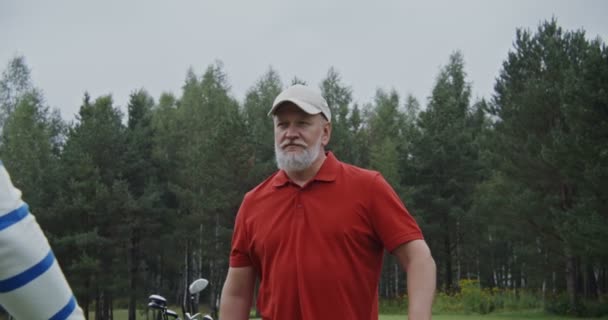 Starszy siwowłosy mężczyzna rozmawia z innym mężczyzną stojąc na polu golfowym — Wideo stockowe