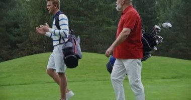 İki adam golf sahasında oyun ekipmanlarıyla yürüyüp birbirleriyle sohbet ediyorlar.