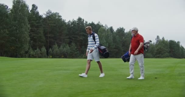 Dos hombres caminan por el campo de golf con bolsas de palos hablando entre sí — Vídeo de stock