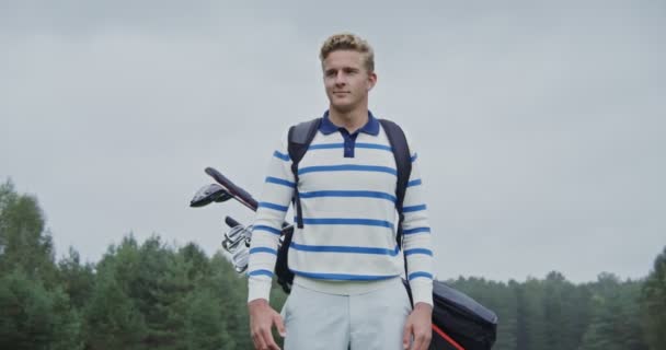 En ung man i sportkläder med golfutrustning bakom ryggen, tittar på kameran — Stockvideo
