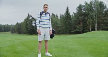 Genç adam kameraya bakıyor ve golf sahasında dikilirken gülümsüyor.