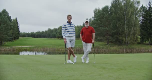 Los hombres están de pie en el campo de golf apoyados en palos de golf y mirando a la cámara — Vídeos de Stock