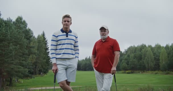 Dois homens estão se apoiando em tacos de golfe no campo de golfe e olhando para a câmera — Vídeo de Stock