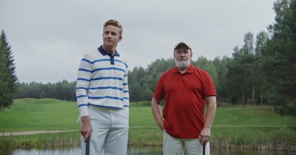 Két férfi golfütőkre támaszkodik a golfpályán, és a kamerába néznek. — Stock videók