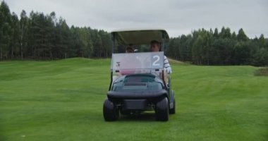 İki adam golf sahasında elektrikli araba kullanıyor, birbirleriyle sohbet ediyorlar.