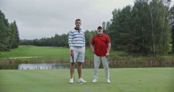 Os homens estão de pé no campo de golfe apoiados em tacos de golfe e olhando para a câmera — Vídeo de Stock