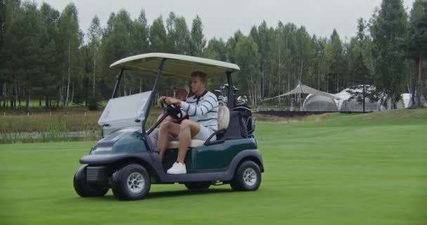 Dois homens estão dirigindo um carro de golfe em um campo de golfe e conversando uns com os outros — Vídeo de Stock