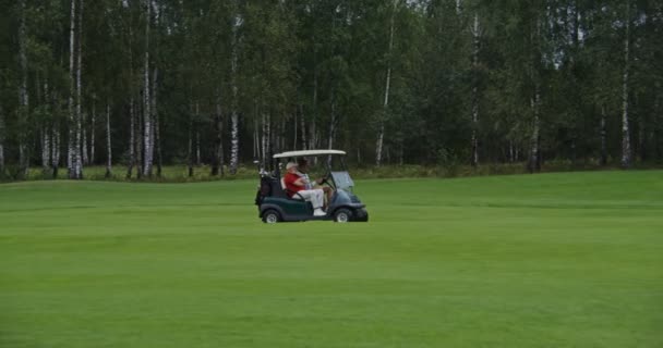 Tampilan samping mobil golf mengemudi melalui lapangan golf dengan dua orang dan peralatan — Stok Video