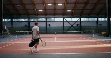 Tenis oynamak üzere olan genç adam yanında bir raket ve bir çanta getiriyor.