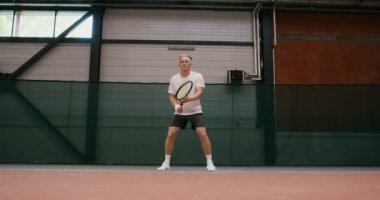 Bir adam tenis oynuyor, raketle topa vuruyor, kapalı alanda oynuyor.