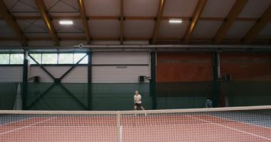 Bir adam tenis oynuyor, raketle topa vuruyor, kapalı alanda oynuyor.