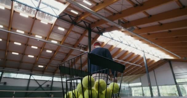 Чоловік грає в теніс сам, беручи м'яч з кошика і кидаючи його через сітку — стокове відео