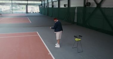Bir adam tek başına tenis oynuyor, bir sepetten topu alıyor ve bir ağın üzerinden atıyor.