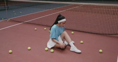 Genç bayan tenisçi tenis kortunun zemininde oturur ve bağcıklarını bağlar.