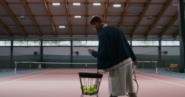 En man spelar tennis ensam, tar en boll från en korg och kastar den över ett nät — Stockvideo