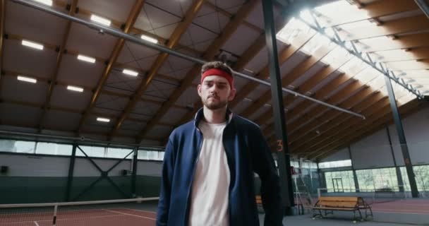 En man står i tennisbana, håller ett tennisracket och en korg full av bollar — Stockvideo