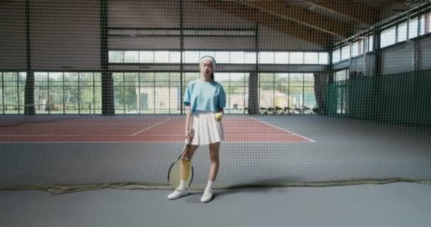 Молодая женщина смотрит прямо в камеру, держа теннисную ракетку и мяч — стоковое видео
