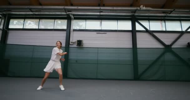 Молодая женщина играет в теннис на крытом теннисном корте, ударяя по мячу ракеткой — стоковое видео