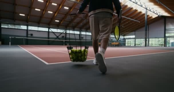 Mężczyzna nosi koszyk pełen piłek tenisowych i rakietę tenisową w rękach. — Wideo stockowe
