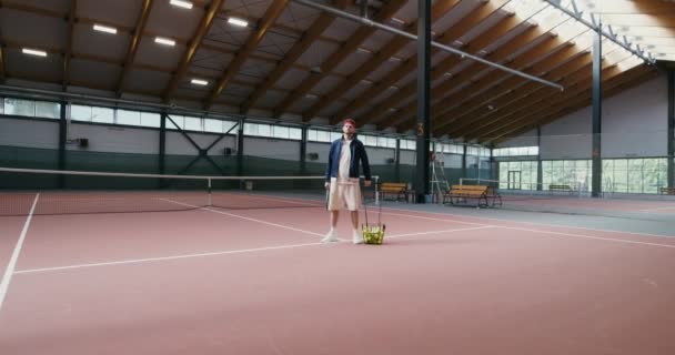 En man står i tennisbana, håller ett tennisracket och en korg full av bollar — Stockvideo