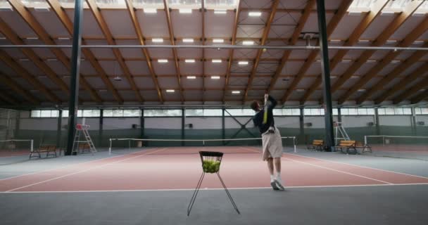 Ein Mann spielt allein Tennis, nimmt einen Ball aus einem Korb und wirft ihn über ein Netz — Stockvideo