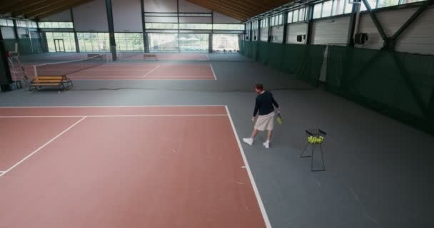 En man spelar tennis ensam, tar en boll från en korg och kastar den över ett nät — Stockvideo