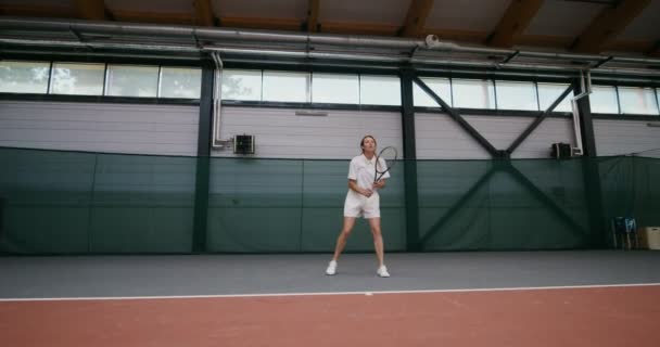 Giovane donna gioca a tennis su un campo da tennis coperto, colpire la palla con una racchetta — Video Stock