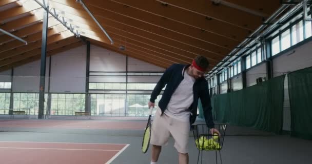 En man spelar tennis ensam, tar en boll från en korg och kastar den över ett nät — Stockvideo