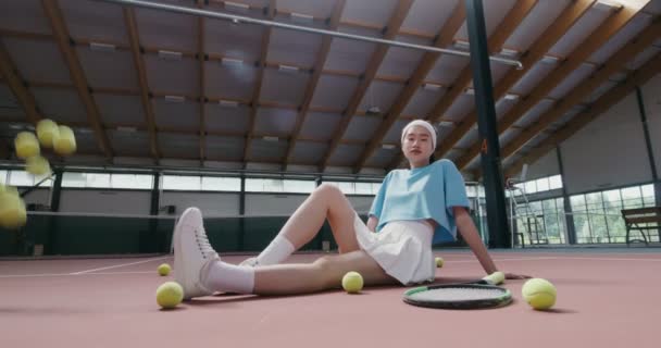 Una mujer joven se sienta en el suelo de la pista de tenis y mira a la cámara — Vídeo de stock