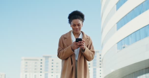 Una mujer de negocios afroamericana sonriendo mientras usa en el teléfono móvil al aire libre — Vídeos de Stock