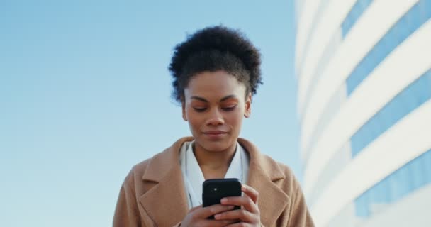 Una mujer de negocios afroamericana sonriendo mientras usa en el teléfono móvil al aire libre — Vídeos de Stock