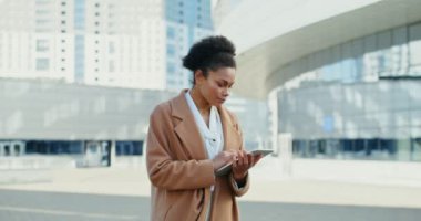 Afro-Amerikalı kadın dışarıda dururken tablet kullanırken gülümsüyor.