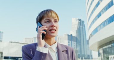 İş elbisesi giymiş bir kadın şehir merkezinde yürürken cep telefonuyla konuşuyor.