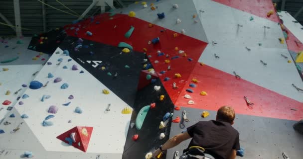 Un joven equipado con arneses de escalada trepa por un muro de escalada — Vídeos de Stock