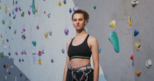Mladá žena dívá do kamery, zatímco stojí před lezeckou stěnou — Stock video