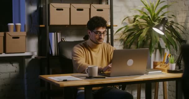 Jovem trabalha em um laptop sentado em uma mesa em um escritório moderno estilo loft — Vídeo de Stock